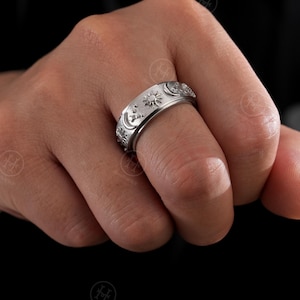 May include: A silver ring featuring engraved celestial motifs such as suns, moons, and stars. The ring is displayed on a finger, highlighting the intricate detailing. The ring's surface has a textured finish, and the design wraps around the band.