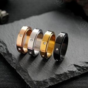 May include: Four rings in rose gold, silver, gold, and black, each with a single, small diamond. The rings are displayed on a dark grey, textured surface. The rings have a modern, minimalist design.