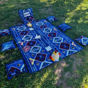 May include: A blue patterned outdoor seating set comprising a large blanket and several matching cushions. The blanket and cushions feature a geometric design in blue, red, and white. The set is arranged on green grass.
