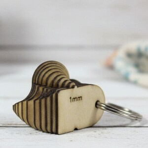 May include: A wooden heart-shaped keychain with a 1mm marking. The keychain is made of layered wood and attached to a silver metal ring. The background includes a succulent plant in a pot and a fabric with floral patterns.