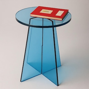 Könnte beinhalten: Ein moderner Beistelltisch mit einer runden, transparenten blauen Tischplatte und einem passenden Gestell. Der Tisch hat ein schwarzes Metallgestell und trägt ein rotes Buch mit goldenem Design. Der Tisch ist etwa 50 cm hoch.