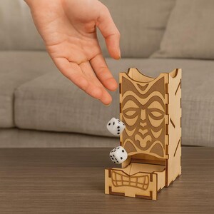 以下が含まれることがあります： 木製のサイコロタワー。ティキの顔のデザイン。黒い点が描かれた白いサイコロが2つ、タワーから落ちています。タワーは明るい色の木材でできており、サイコロが転がり出るスロットがあります。