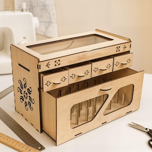 Puede incluir: Un organizador de costura de madera con múltiples cajones y compartimentos. El organizador tiene una parte superior transparente y cortes florales decorativos. Los cajones tienen tiradores decorativos. Diseñado para guardar hilos y suministros de costura.