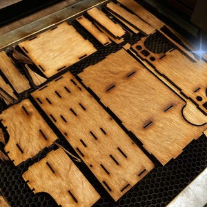 May include: Laser-cut wooden pieces arranged on a honeycomb surface. The pieces are various shapes and sizes, with slots and cutouts. The wood has a warm, natural tone. The laser is actively cutting a piece, creating a bright blue light.