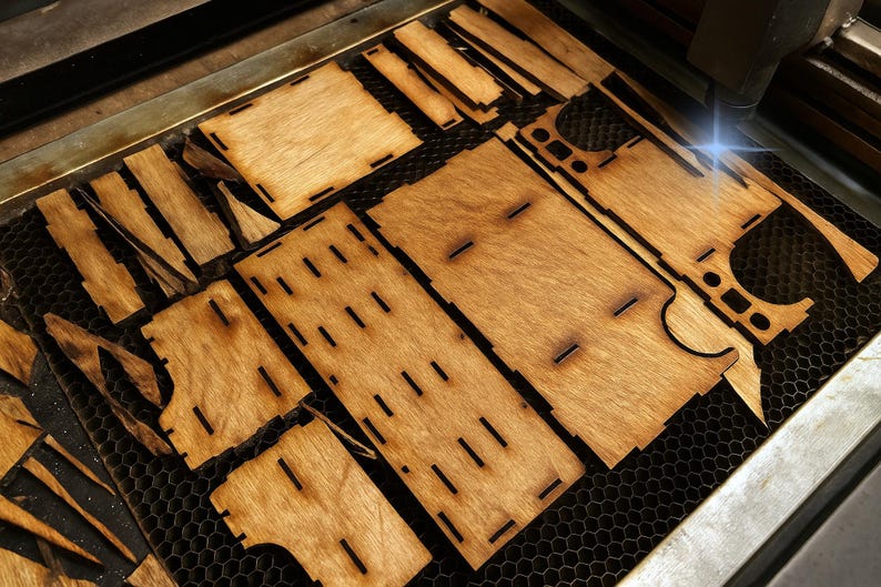 May include: Laser-cut wooden pieces arranged on a honeycomb surface. The pieces are various shapes and sizes, with slots and cutouts. The wood has a warm, natural tone. The laser is actively cutting a piece, creating a bright blue light.