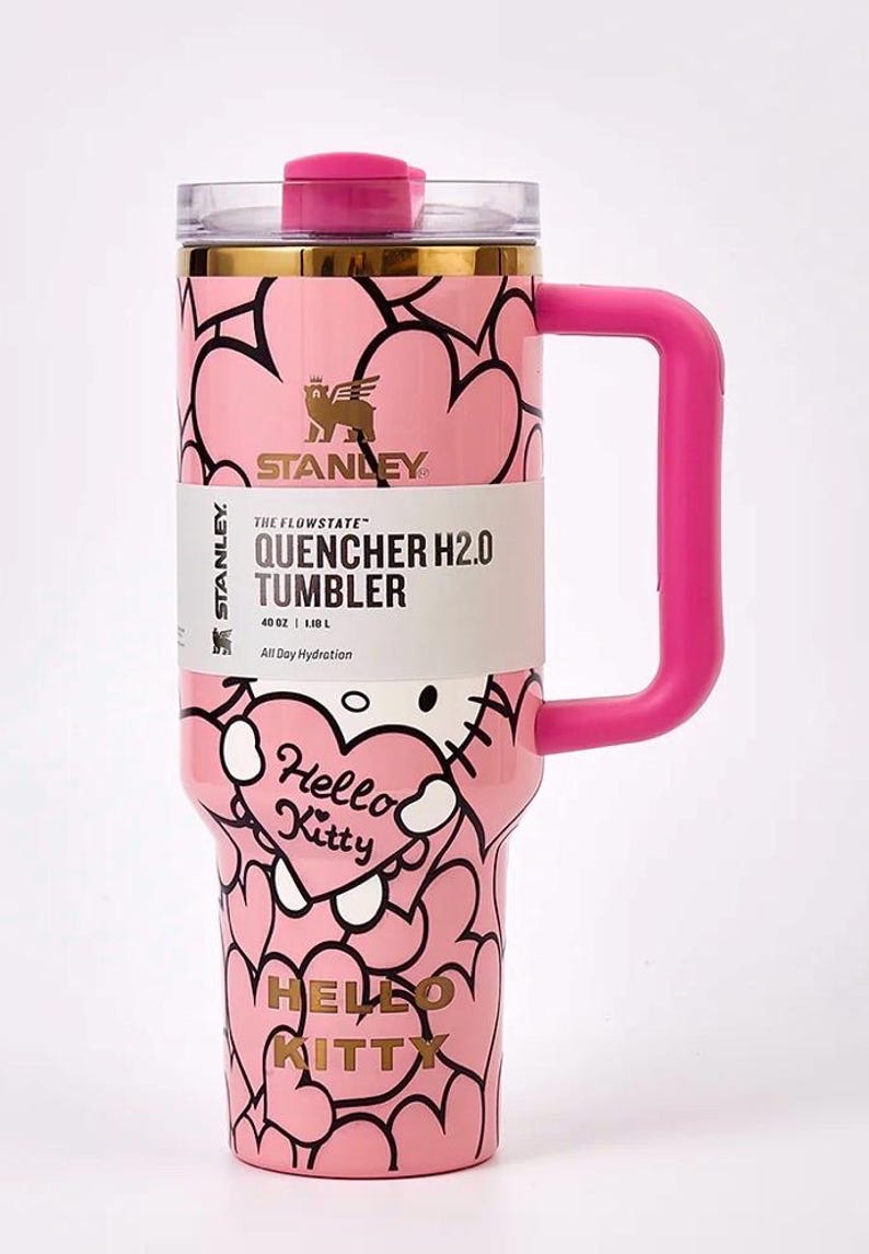 May include: Pink Stanley Quencher H2.0 tumbler with a Hello Kitty design. The tumbler has a pink handle and lid, and features a heart pattern. The tumbler holds 40 oz (1.18 L) and has the text "Hello Kitty" on the front.