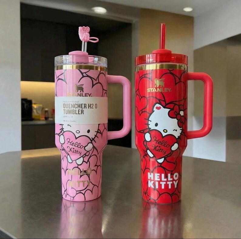 May include: Two Stanley Quencher H2.0 tumblers, one pink and one red, featuring Hello Kitty designs. The pink tumbler has a heart pattern and the text "Hello Kitty". The red tumbler has a Hello Kitty graphic holding a heart. Both have handles and straws.