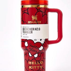 May include: Red Stanley Quencher H2.0 tumbler with a Hello Kitty design. The tumbler features a red handle, lid, and a gold rim. The body is covered in black heart outlines and Hello Kitty graphics. The tumbler has a 40 oz capacity.