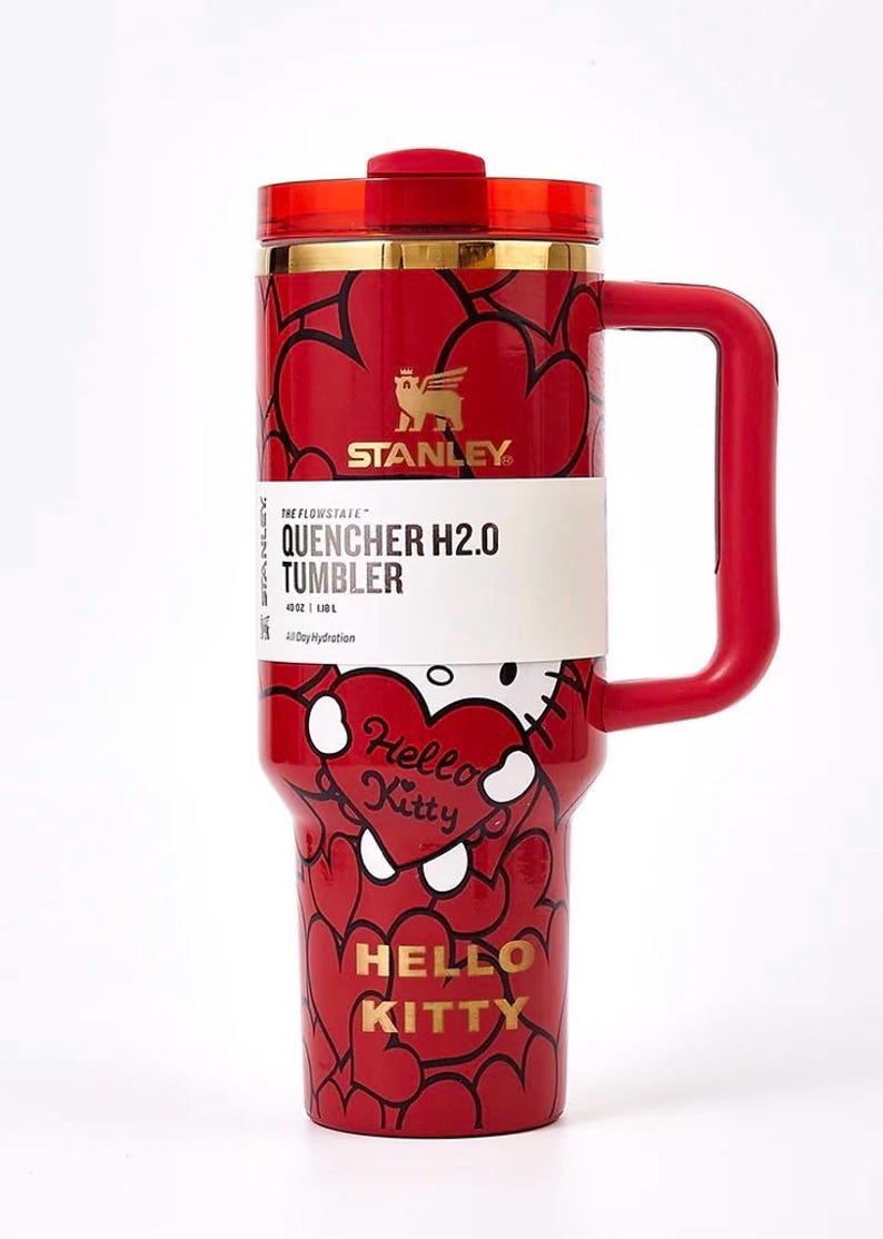 May include: Red Stanley Quencher H2.0 tumbler with a Hello Kitty design. The tumbler features a red handle, lid, and a gold rim. The body is covered in black heart outlines and Hello Kitty graphics. The tumbler has a 40 oz capacity.