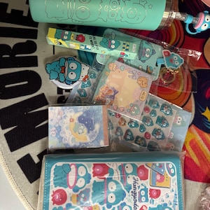 May include: A collection of stationery and accessories featuring a cartoon character in shades of blue and teal. Items include a tumbler, stickers, a pencil case, and notepads. The pencil case has the character's name, "Hangyodon", printed on it.