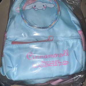 May include: A light blue backpack featuring a circular clear window with a Cinnamoroll character graphic. The front pocket has a pink zip and the text Cinnamoroll, along with the phrase Welcome to my room! I love doing the things I love. What makes you happy? The bag includes pink side mesh pockets and pink straps.