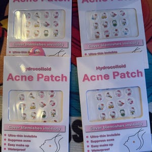 May include: Four packages of hydrocolloid acne patches. Each package has a white background with the words "Hydrocolloid Acne Patch" in pink and white. The patches are round and feature a cartoon cat design. The text "Cover blemishes invisibly" is also visible.