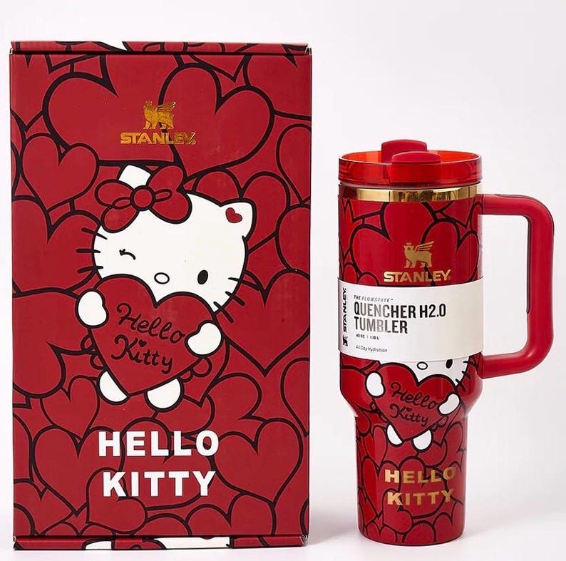 May include: A red Stanley Quencher H2.0 tumbler and matching box featuring Hello Kitty graphics. The tumbler has a handle and a gold band. The box and tumbler are covered in red hearts. The tumbler has the text "Hello Kitty" and "Quencher H2.0 Tumbler".