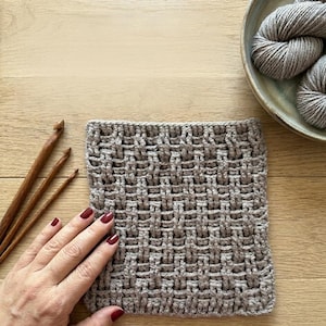 May include: A close-up of a crocheted square in a basketweave pattern, in a neutral beige color. The square is on a wooden surface, with three wooden crochet hooks to the left and a bowl of gray yarn in the upper right corner.