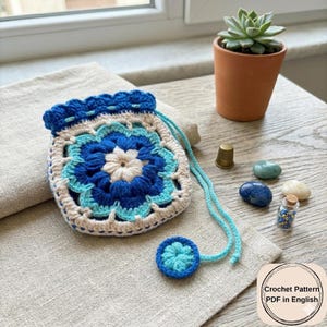May include: A handmade crochet pouch in shades of blue, cream, and white, featuring a floral design. The drawstring bag is displayed on a linen surface, with a small matching button. A small terracotta pot with a succulent plant is in the background.