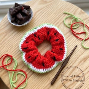 May include: A handmade, crocheted watermelon-shaped scrunchie with red, white, green, and black details. The scrunchie is on a wooden surface, with a crochet hook, yarn, and a small bowl of chocolate-covered treats. The text "Crochet Pattern PDF in English" is visible.