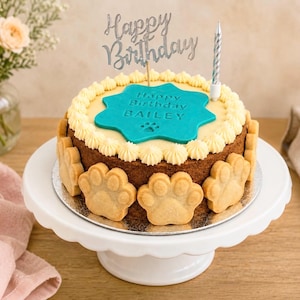 May include: A round cake with brown sides decorated with light brown paw-shaped cookies, topped with cream frosting, a teal star-shaped plaque reading Happy Birthday BAILEY with a paw print, a silver Happy Birthday cake topper, and a silver candle.
