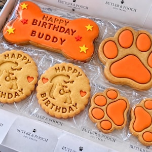 May include: A white box containing dog treats including one large orange bone-shaped cookie with the text HAPPY BIRTHDAY BUDDY and yellow stars, two round cookies with HAPPY BIRTHDAY text, and three orange paw-shaped cookies with Butler & Pooch branding.