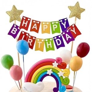 May include: A vibrant cake decoration featuring a rainbow, balloons, and a banner spelling "HAPPY BIRTHDAY." The topper includes gold glitter stars and is designed for a celebratory occasion. The balloons are in various colours.