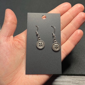 May include: Silver-toned button earrings with four holes in each button, hanging from a simple hook. The earrings are displayed on a black card, held in a hand.