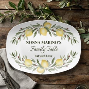 May include: White oval serving platter with a lemon and olive branch design. The platter has the text "NONNA MARINO'S Family Table Eat with Love". The platter is on a wooden table with silverware and a napkin.
