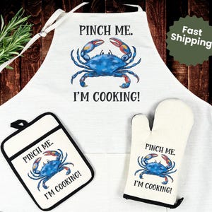 May include: White apron, oven mitt, and pot holder set with a blue crab design. The apron and accessories feature the text "PINCH ME. I'M COOKING!" A sprig of rosemary is visible on a wooden surface.