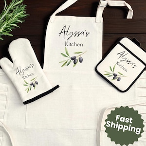 May include: A white kitchen apron, oven mitt, and pot holder set. Each item features the text "Alyssa's Kitchen" with an olive branch design. The apron has a pocket and ruffled trim. The oven mitt and pot holder have black trim.