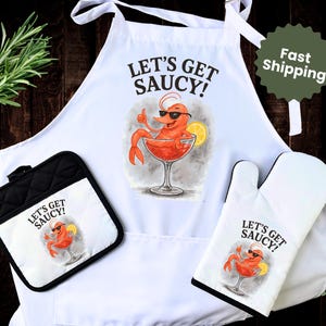 May include: A white apron, pot holder, and oven mitt set. Each item features a cartoon shrimp in a cocktail glass with sunglasses and the text "Let's Get Saucy!". The pot holder is black and white.