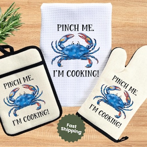 May include: A kitchen set featuring a pot holder, oven mitt, and dish towel. Each item is off-white with a black border and features a blue crab illustration with the text "PINCH ME. I'M COOKING!".