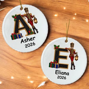 May include: Two round white ceramic ornaments featuring a black letter A or E, a skeleton nutcracker figure holding a lantern, holly berries, and wrapped gift boxes. The text Asher 2026 or Eliana 2026 is printed below each letter.