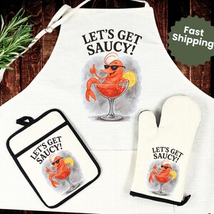 May include: White kitchen apron, pot holder, and oven mitt set. Each item features a cartoon shrimp in a cocktail glass with the text "LET'S GET SAUCY!". The apron has a neck strap and waist ties.