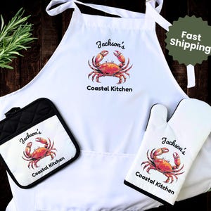 May include: A white apron, oven mitt, and pot holder set featuring a watercolor crab design. The text "Jackson's Coastal Kitchen" is printed on each item. The apron has tie straps, and the pot holder and oven mitt are black and white.