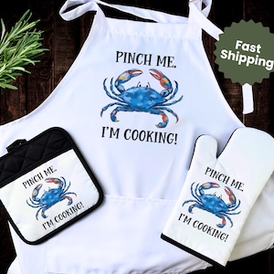May include: White apron, oven mitt, and pot holder set featuring a blue crab design with the text "PINCH ME. I'M COOKING!". The apron has ties, and the pot holder is black and white.