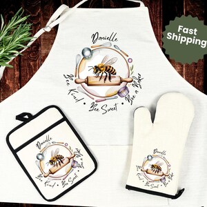 May include: A white apron, pot holder, and oven mitt set featuring a bee-themed design. The apron and accessories display a bee illustration with the words "Bee Kind, Bee Sweet, Bee a Baker". The set is perfect for baking.