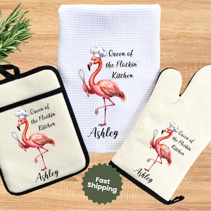 May include: A kitchen set featuring a flamingo design. The set includes a pot holder, oven mitt, and a kitchen towel. Each item has a pink flamingo wearing a chef's hat, holding a whisk, and the text "Queen of the Flockin' Kitchen" and the name "Ashley".