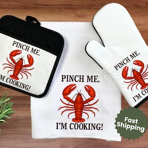 May include: A kitchen set featuring a black pot holder, white oven mitt, and a white kitchen towel. Each item has a red lobster graphic and the text "PINCH ME. I'M COOKING!" printed on them. The set is designed for cooking.