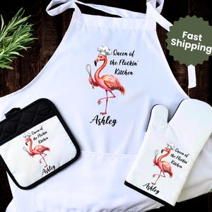 May include: A white kitchen apron, pot holder, and oven mitt set. Each item features a pink flamingo wearing a chef's hat and holding a whisk. The text "Queen of the Flockin' Kitchen" and the name "Ashley" are also printed on each item.