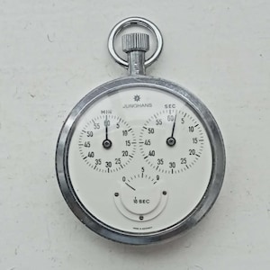 May include: A silver-toned Junghans stopwatch with a round metal case and a white face. The dial features two main sub-dials labeled MIN and SEC, and a smaller bottom sub-dial labeled 1/10 SEC. The text JUNGHANS and MADE IN GERMANY are printed on the face.