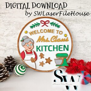 以下が含まれることがあります： 白い背景に「WELCOME TO Mrs. Claus KITCHEN」という文字が書かれた円形の木製看板。ミセス・クロースのカラフルなイラスト、緑色のスタンドミキサー、ジンジャーブレッドクッキー、お祝いの松の枝がデザインされています。