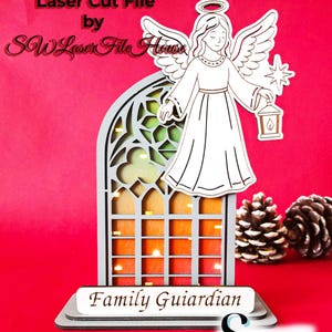 Può includere: Design in legno tagliato al laser di un angelo e di una vetrata con la scritta "Family Guardian". L'angelo tiene in mano una lanterna e una stella. Sfondo rosso con pigne. Testo "Laser Cut File by SW Laser File House".