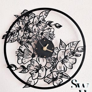 May include: Black and white laser cut wall clock with a floral design. The clock face is black with gold hands. The clock is surrounded by a circle of flowers.