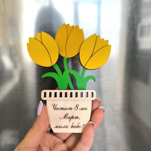 May include: A wooden decorative magnet featuring three bright yellow tulips with green stems and leaves emerging from a light-colored pot. The pot displays the text "Честит 8-ми Март, мила бабо!" in a dark cursive script, with a row of vertical cutouts.
