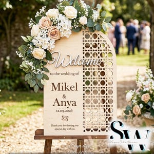 Puede incluir: Un letrero de bienvenida de boda de madera arqueado con un patrón geométrico cortado con láser en el lado derecho y un arreglo floral de rosas crema y eucalipto en la parte superior izquierda. El letrero muestra el texto Welcome to the wedding of Mikel & Anya 12.09.2026.
