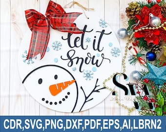 May include: A round white wooden sign featuring a snowman face, a red and black plaid bow, and the text "let it snow" in black script. The design includes small blue snowflake cutouts and is presented as a digital download file for laser cutting.