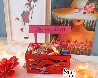 May include: A red wooden crate labeled Kissing Booth with a cutout of red lips and small pink hearts on the front. The crate is filled with various colorful wrapped candies and features a handle with the text Kissing Booth in black script.