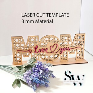 May include: A laser-cut wooden sign featuring the word MOM with a cane webbing pattern, overlaid with the pink script text I Love You. The sign is designed for 3 mm material and includes file formats SVG, CDR, AI, DXF, EPS, PDF, PNG, and Lightburn.