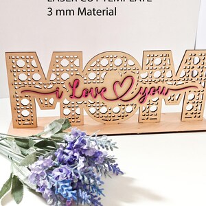 May include: A laser cut wooden sign featuring the word MOM with a cane webbing pattern, layered with a pink script text reading I Love You. The sign is designed for 3 mm material and includes file formats SVG, CDR, AI, DXF, EPS, PDF, PNG, and Lightburn.