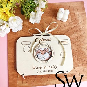 May include: A light-colored wooden camera-shaped photo frame featuring the text Captured, Forever Starts Here, Mark & Lilly, and July 12th 2026. The frame includes a circular center cutout displaying a group photograph and is tied with a cream ribbon.