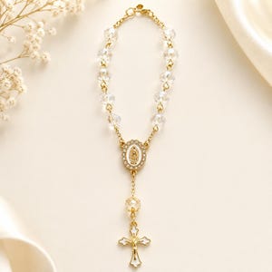 May include: A gold-toned rosary bracelet featuring clear faceted crystal beads, a central oval charm with a white enamel inlay depicting a figure, and a gold-toned cross pendant with white enamel detailing, all connected by a delicate gold-toned chain.