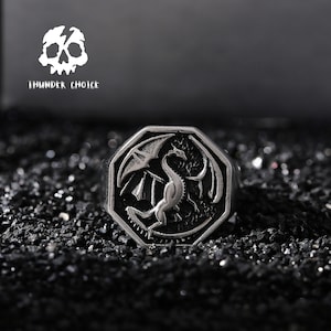 Könnte beinhalten: Ein silberner Ring mit einem achteckigen Design, das einen detaillierten Drachen mit ausgebreiteten Flügeln zeigt. Der Ring befindet sich auf einem schwarzen Hintergrund.